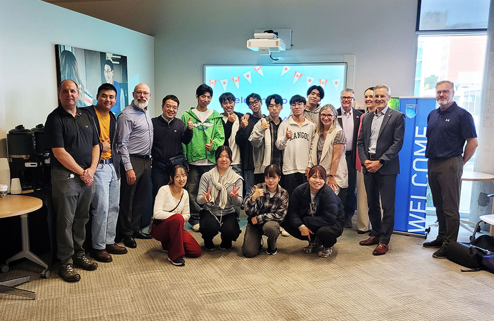 Group photo of 17 people in an indoor setting with office furniture. Some are standing, others kneeling or sitting. A projector screen in the background shows colorful bunting flags, and a 'WELCOME' banner is partially visible to the right.