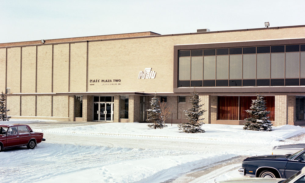 NAIT Plaza Two in winter, a beige brick two-story building with flat roof, recessed glass entrance, and a large section of dark-tinted windows. Snow and evergreen trees in front, cars parked nearby.