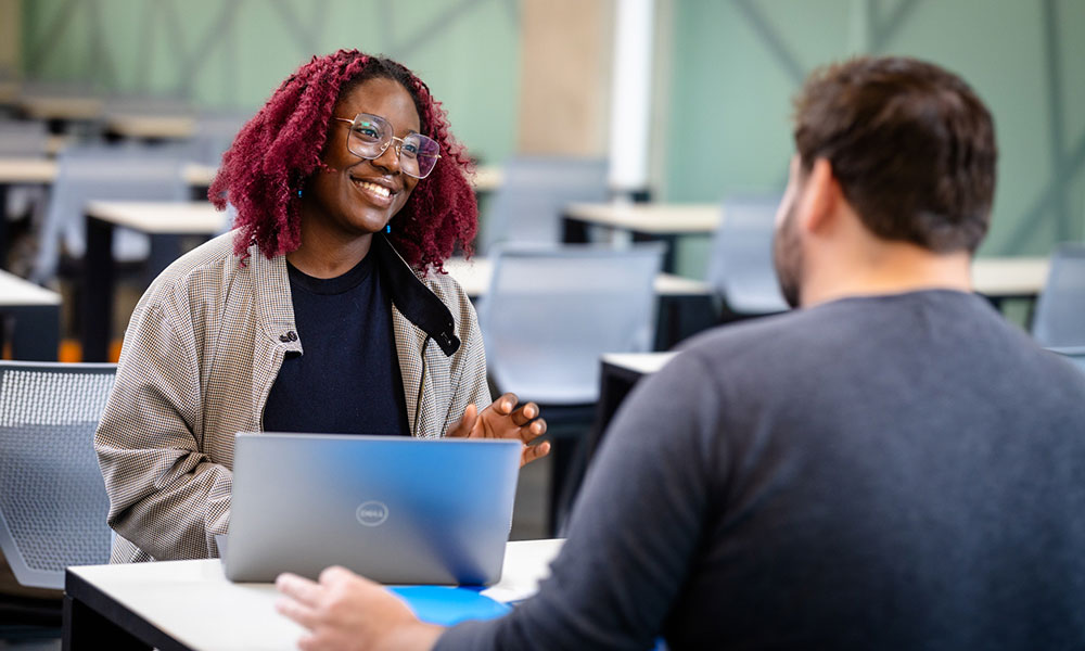 Two people sit across from each other at a table in a large room with rows of desks; one is using a laptop.