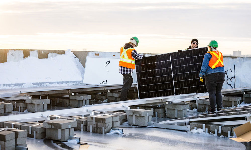 two people in green hard hats and orange safety vests carry a black solar panel on a rooftop