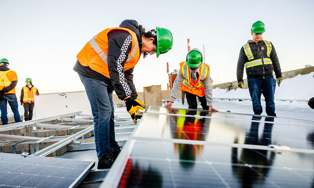 a woman in a green hard hat and an orange safety vest bends over a solar panel being installed on a rooftop
