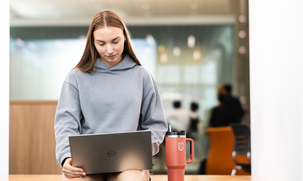 Person sitting indoors with a laptop on their lap and a large insulated mug beside them.