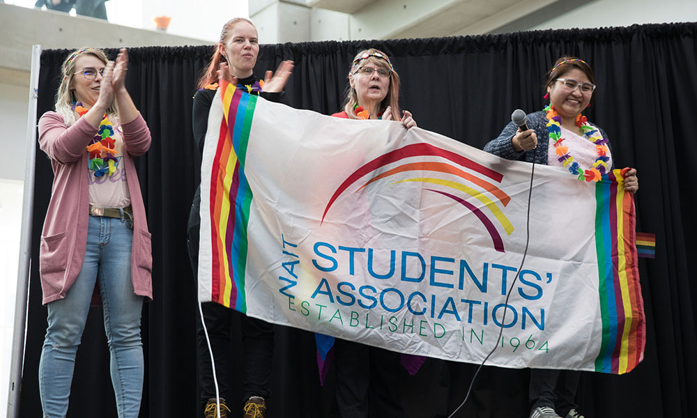 A group of individuals standing on a stage holding a large NAIT Students’ Association banner with rainbow-themed designs, while wearing colourful leis and standing in front of a black curtain.