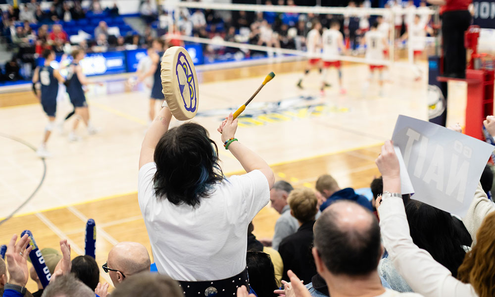A crowd watches an indoor volleyball match as a spectator in the foreground raises a small drum and mallet, with players and the court visible in the background.