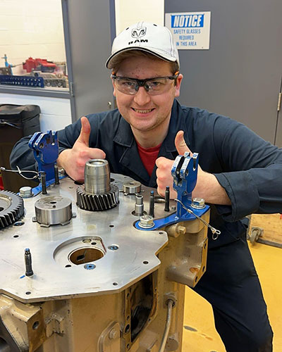 a young man in a white baseball cap and safety glasses crouches by a piece of machinery and gives a thumbs up while smiling