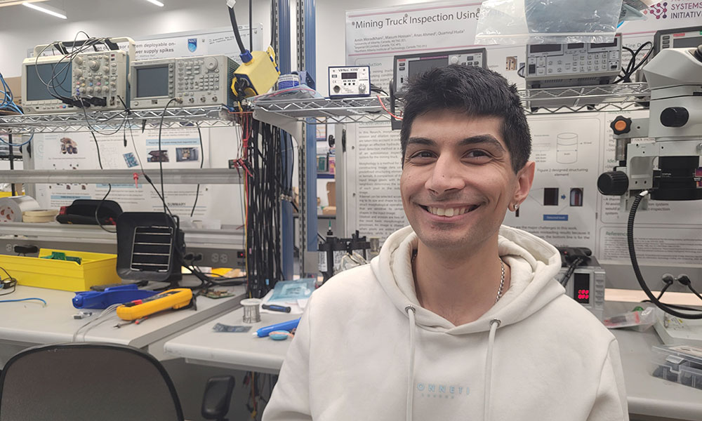 Person in a white hoodie sitting at an electronics workbench with and electronics various tools. Shelves above hold equipment and cables, and technical posters are displayed on the wall in the background.