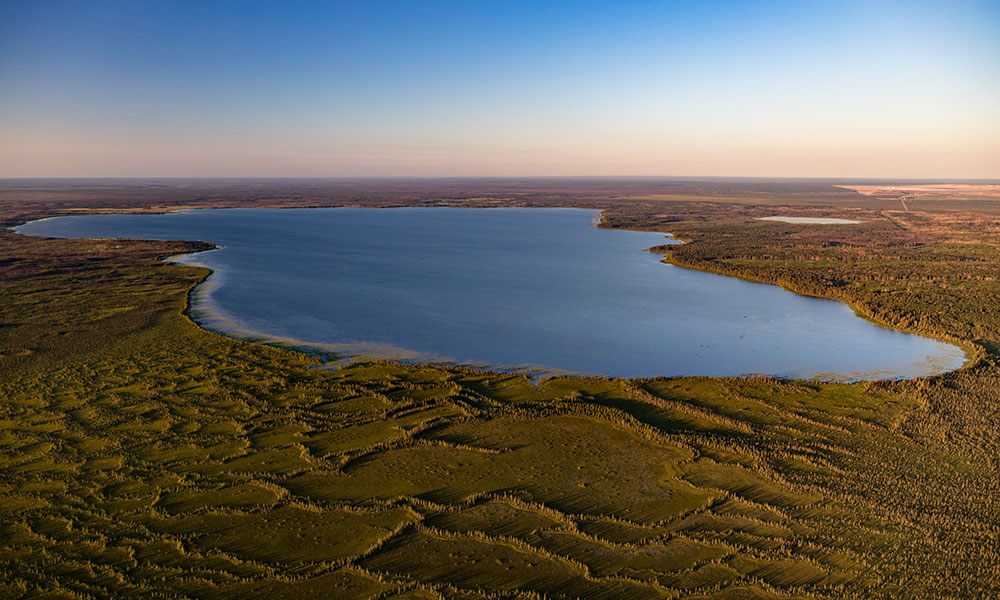 Aerial view of a large lake surrounded by expansive green, gently rolling terrain. The horizon is visible under a clear blue sky that fades to a warm hue near the horizon, with no signs of human activity.