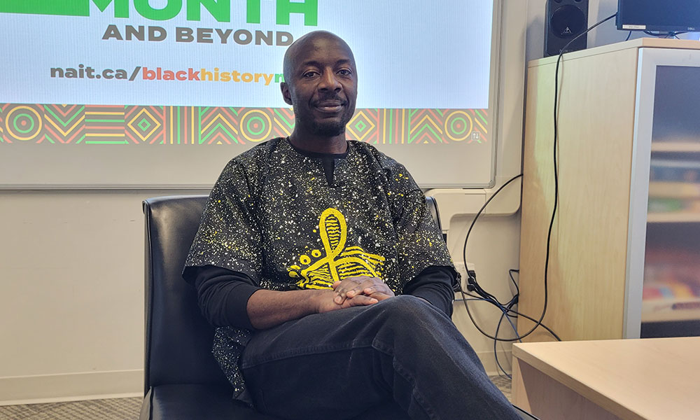 A person seated in a chair wearing a patterned shirt with an ankh design, positioned in front of a screen displaying “Black History Month and Beyond.”