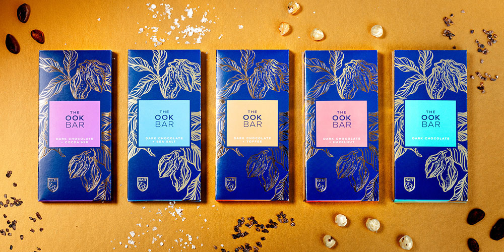 Five blue‑and‑gold “The Ook Bar” dark chocolate bars displayed side by side on a golden background with scattered cocoa nibs and ingredients.