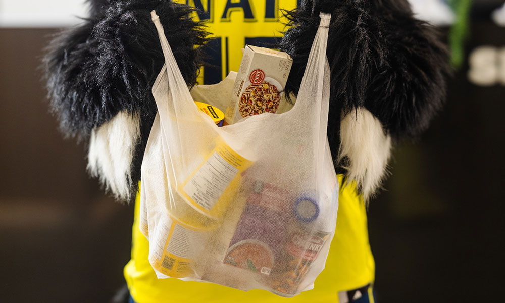 A person in a black-and-white furry costume wearing a bright yellow shirt holds a clear plastic grocery bag filled with packaged food items, including cans and a box of pasta.
