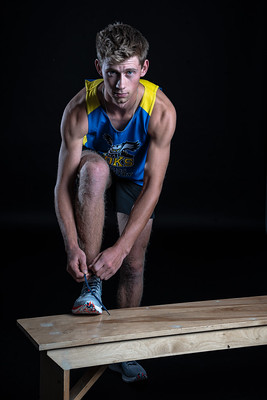 Athlete wearing a blue and yellow tank top sits on a wooden bench, tying their shoe.