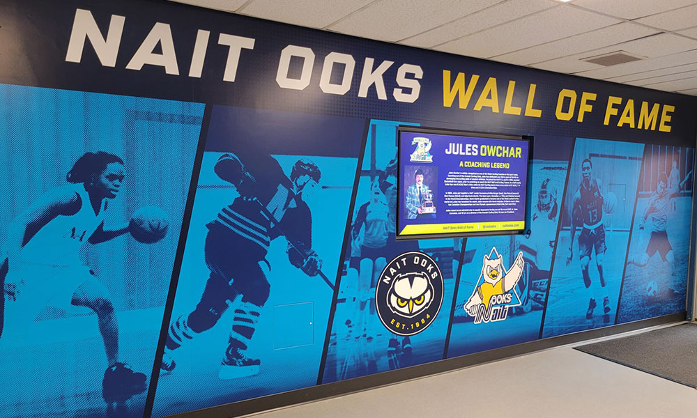 Display wall titled 'NAIT Ooks Wall of Fame' featuring large images of athletes from sports like basketball and hockey. Central section highlights coach Jules Owchar with text about his achievements. NAIT Ooks logo and mascot also visible.