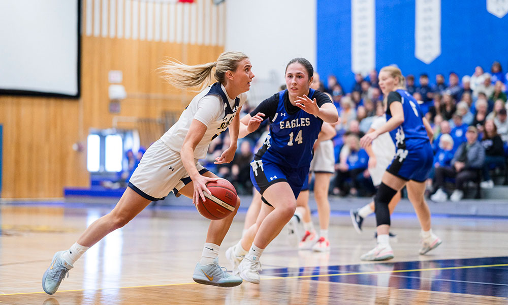 Two basketball players contest the ball during an indoor game on a hardwood court, with teammates and spectators visible in the background.
