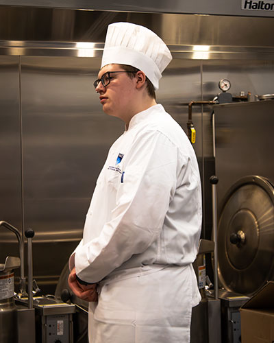 a young man in stands in a commercial kitchen wearing a chef's jacket and hat