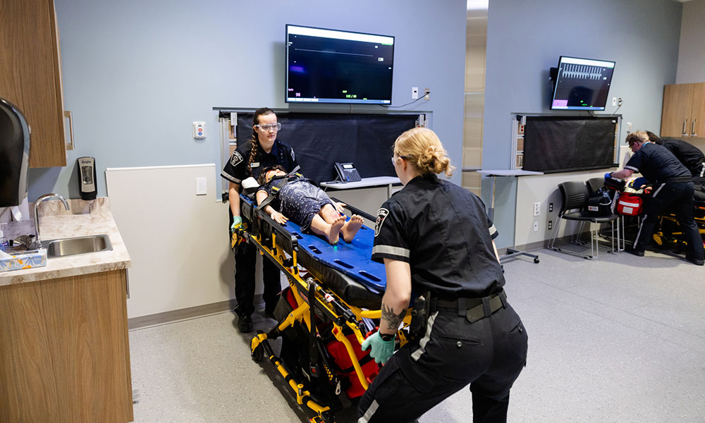 Three paramedics attending to a patient on a stretcher in a medical emergency room, with monitors displaying medical data and various equipment visible.