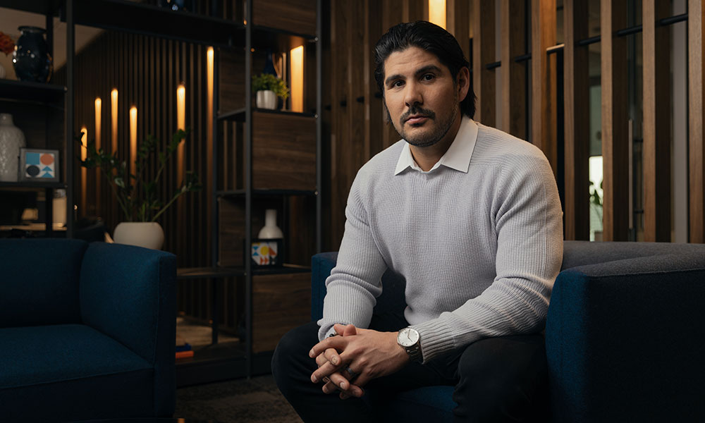 Person sits on a blue chair in a modern, warmly lit office lounge with wood shelving and decorative accents in the background.