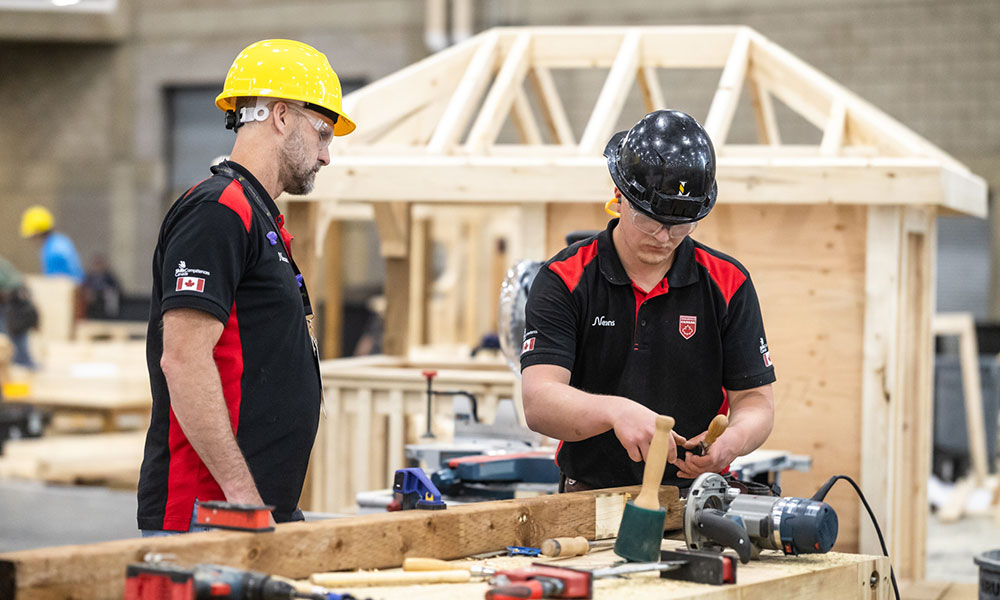 Two people in safety helmets working at a carpentry station with hand tools, assembling a wooden structure in a large indoor workshop.