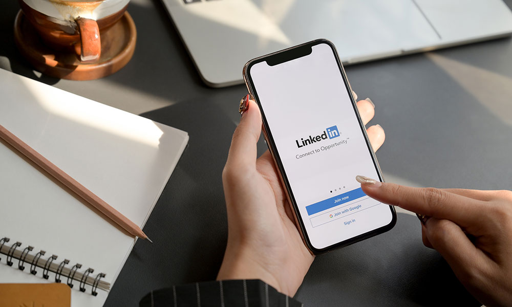 Close-up of a person's hands using the LinkedIn app on a mobile phone, with a laptop and notebook in the background.