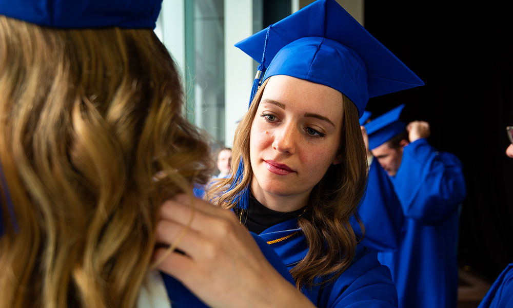 Group of people in blue graduation gowns and caps. One person, with a blurred face, is adjusting the ribbon on another person's gown.