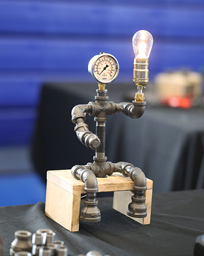 A small lamp sculpture made from metal pipes and fittings sits on a wooden block. The design resembles a seated figure, with a pressure gauge as the head and a glowing light bulb held up like an arm.