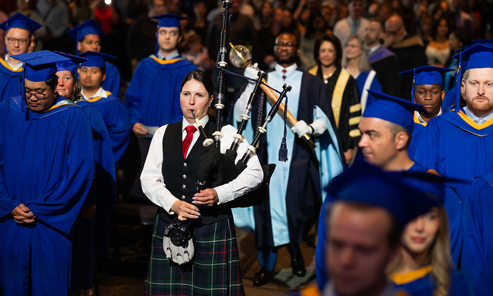 Graduation ceremony with graduates in blue gowns and caps. A bagpiper in traditional Scottish attire, including a kilt, leads the procession. Individuals in academic regalia follow behind, with an audience in the background.