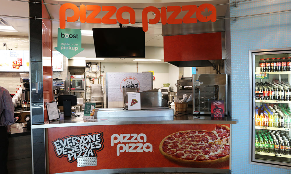 Pizza restaurant counter labeled 'pizza pizza' with a mobile order pickup sign 'boost'; counter includes a menu, promotional materials, and a cash register. A refrigerator with beverages is on the right, and the front of the counter reads 'EVERYONE DESERVES PIZZA' with an image of a pepperoni pizza.