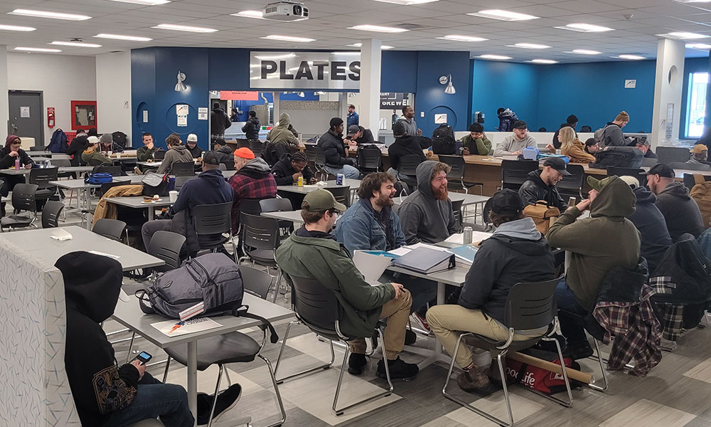 Students are seated at tables in a cafeteria-style space called “PLATES,” studying and socializing, with notebooks and bags on the tables.