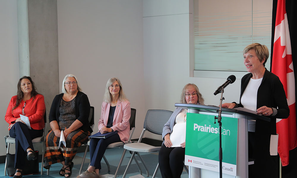 Five people in a room, four seated and one standing at a podium labeled 'PrairiesCan' with Government of Canada logos. A Canadian flag is visible in the background.