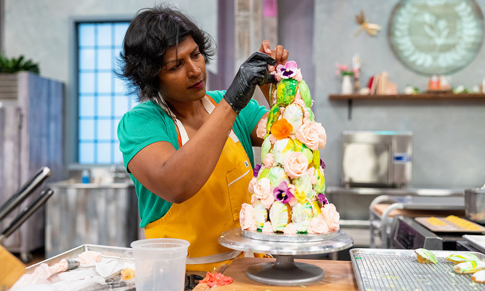 Photo courtesy of Food Network a woman in a commercial kitchen assembles a very tall and artistic cake decorated with many colours and flowers