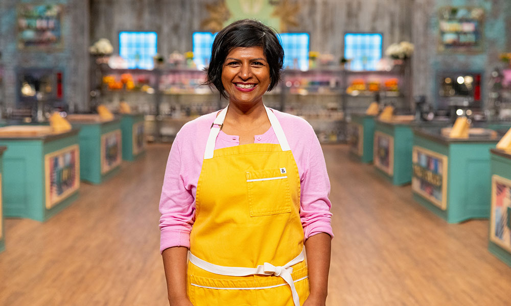 Photo courtesy of Food Network a woman in a pink sweater and yellow apron smiles for a photo on the set of a cooking show. there are blurred shelves behind her and green workstations on either side of the room.