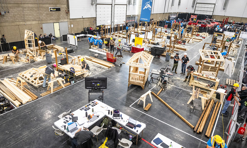 Large indoor carpentry workshop with multiple participants building wooden structures at individual stations across a spacious hall.