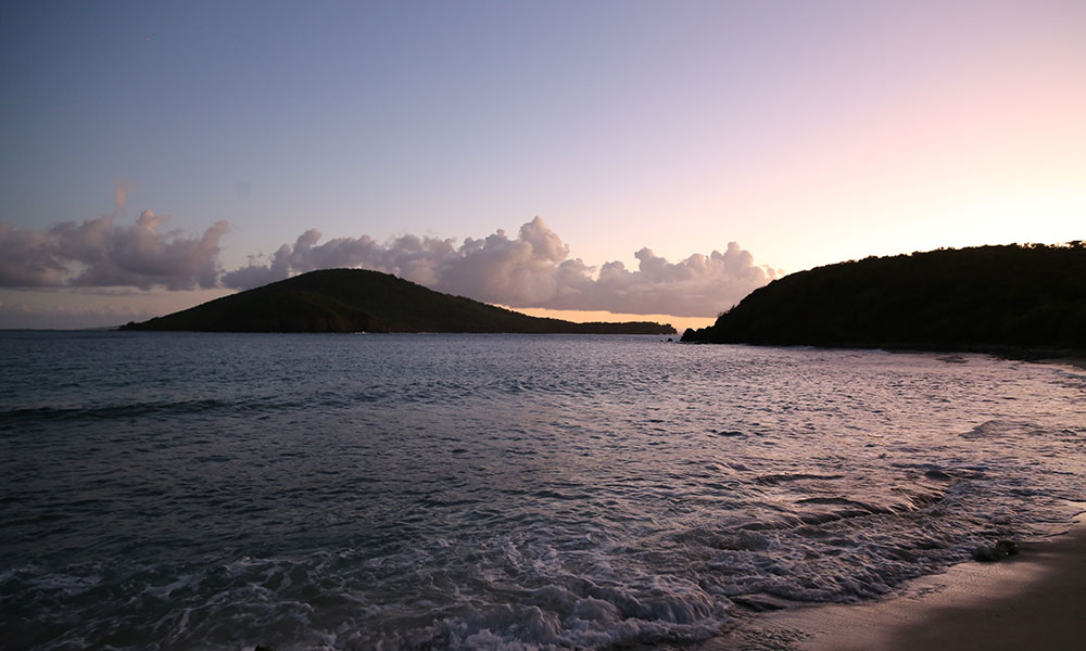 A serene beach scene at sunset or sunrise with a gradient sky transitioning from pink to purple. Gentle ocean waves meet the shore, and two distant, vegetation-covered landmasses are visible—one larger on the left and a smaller one on the right. Scattered clouds float above the horizon.