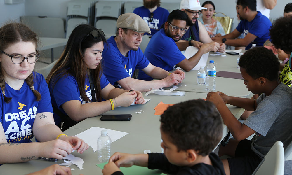 A group of people sitting around a table engaged in a crafting activity. Most are wearing blue shirts with the text 'DREAM ACT ACHIEVE' and 'BETTER FUTURE.' The table has water bottles and pieces of paper on it.
