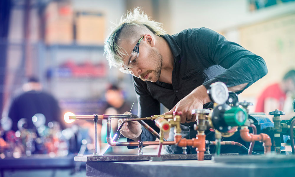 A person with light-colored undercut hair, wearing a dark long-sleeve shirt, uses a blowtorch at a workbench in a workshop or lab. The bench is cluttered with tools, pipes, pressure gauges, and valves. The background is blurred, showing other people and equipment, suggesting a busy environment.
