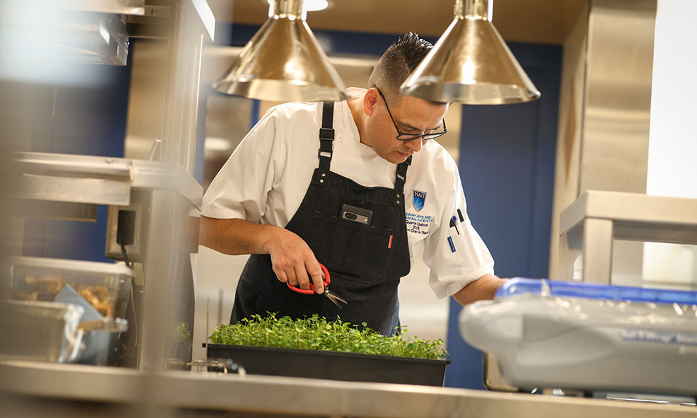 a chef cuts ingredients from a tray of microgreens in a commercial kitchen