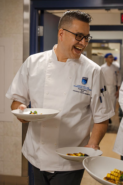 a chef smiles to someone off camera while he carries a plate of food in each hand