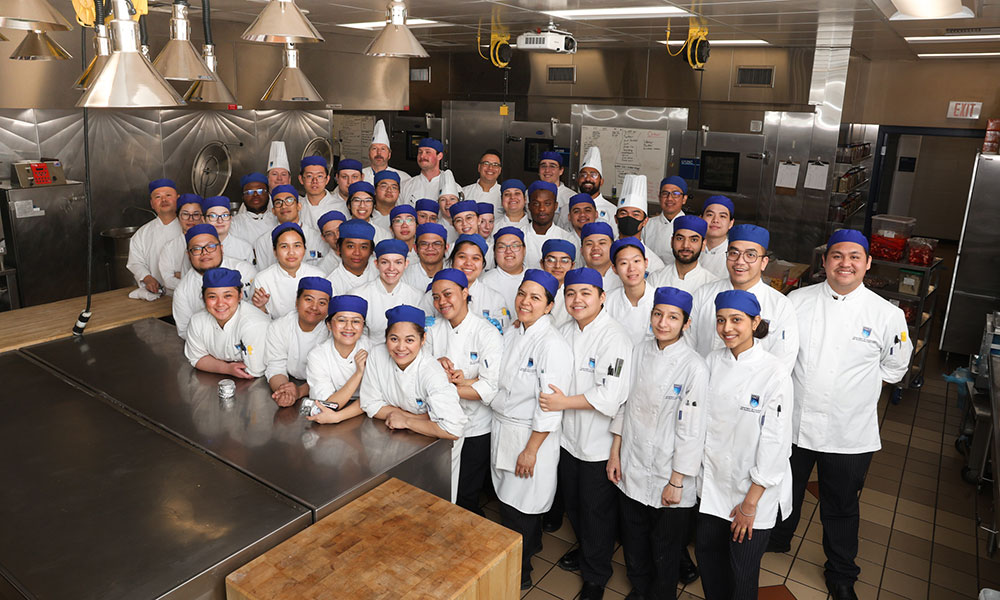 a group photo of culinary students in chef's whites and blue caps in a commercial kitchen