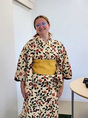 Individual wearing a cream-colored floral kimono with a yellow obi, standing indoors near a white wall and air conditioning unit. A round table with objects is partially visible in the foreground.