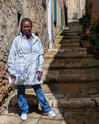A person in a light jacket, blue jeans, and white shoes stands on a stone-paved alleyway with steps. The alley is flanked by stone walls, one side featuring blue-shuttered windows and potted plants.