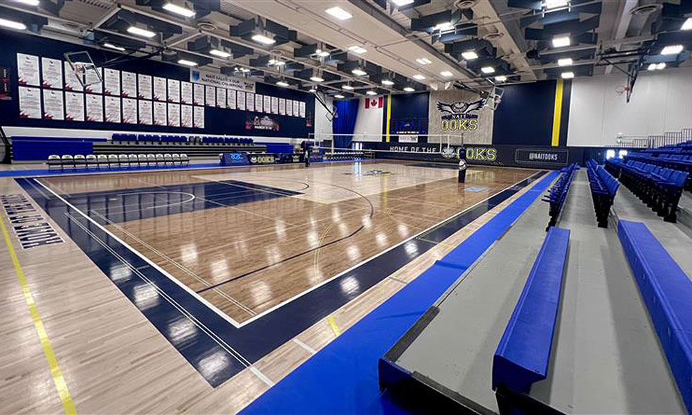 A renovated gymnasium viewed from the corner, showing a polished court, blue bleachers, and banners lining the far wall.