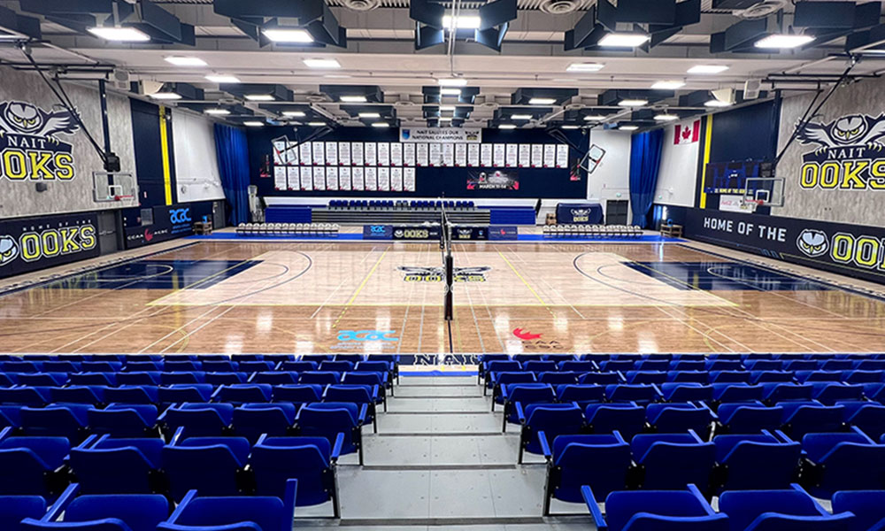 A wide view of an empty, renovated indoor gymnasium with a polished court, blue seating, banners, and team branding on the walls.