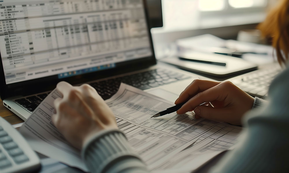 Hands reviewing printed financial documents with a pen in front of a laptop displaying spreadsheets.
