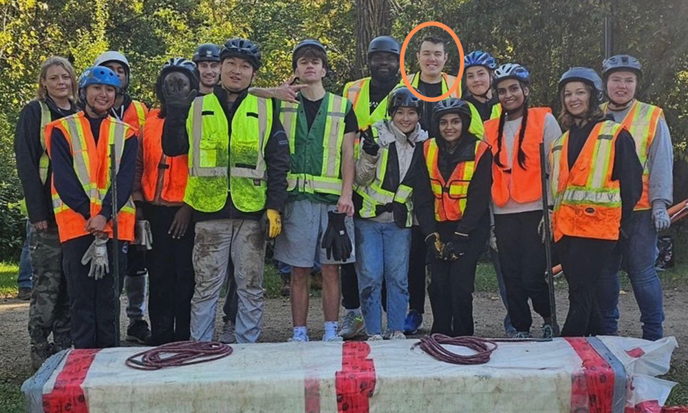 A group of people wearing safety vests and helmets stand outdoors around a large object wrapped in white material with red tape and ropes. One individual in the back row, second from the right, is circled in orange.
