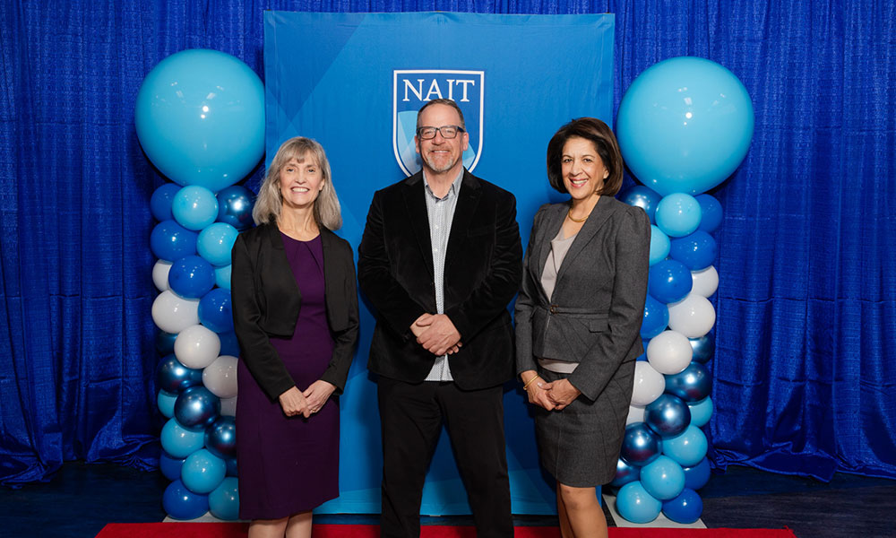 two women pose for a photo with a man standing between them. they are wearing business and formal attire. behind them is a blue background and blue balloons.