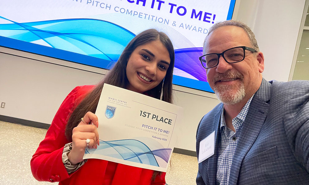 A person holding a “1st Place – Pitch It to Me!” award stands beside another person in front of a large event screen with blue wave graphics.