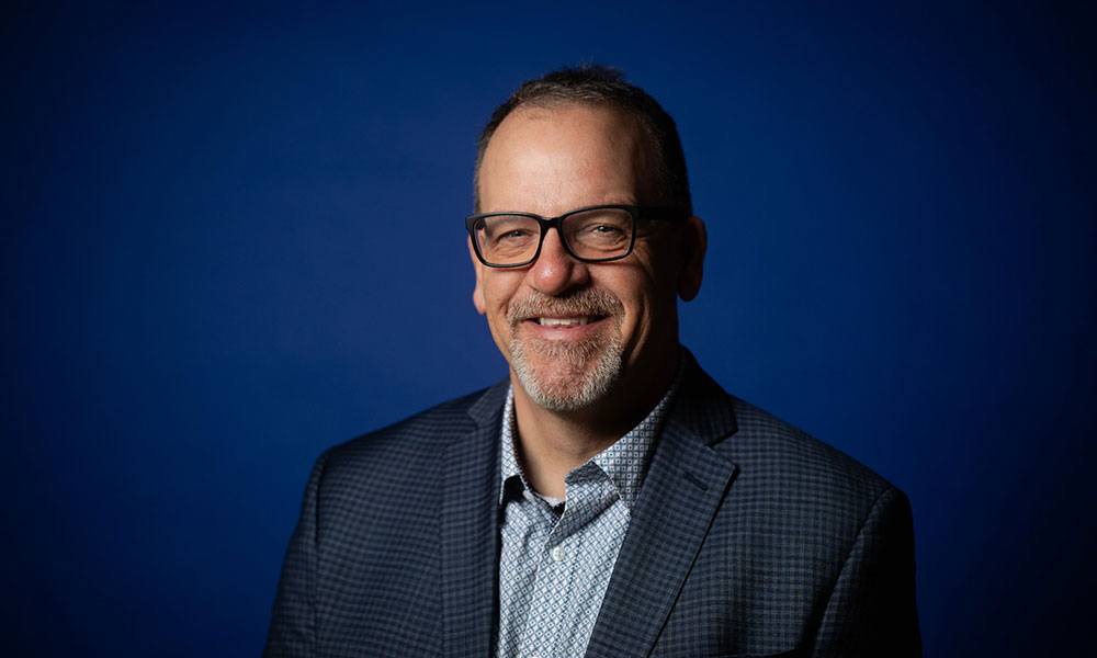 A person wearing a patterned shirt and checkered blazer is photographed against a solid blue background.