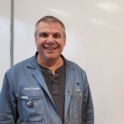 A person wearing a blue lab coat with visible text “Rob Csolle” and a patch labeled “NAIT” is standing in front of a whiteboard, with pens in the coat pocket and a button pinned to the coat.