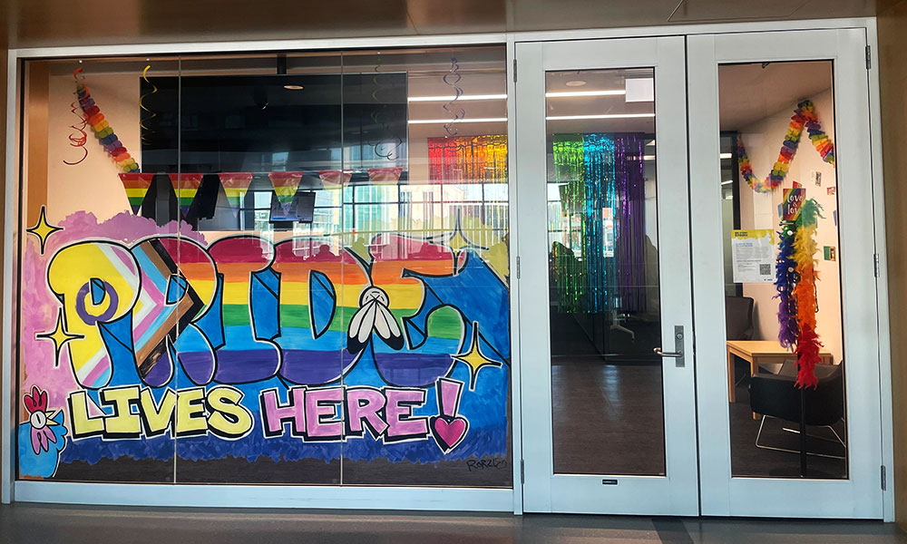 A brightly decorated glass storefront featuring a large, colourful mural that reads “PRIDE LIVES HERE!” in bold, graffiti‑style lettering, surrounded by rainbow‑themed decorations and streamers visible inside the room.