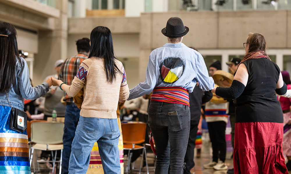 People stand in a circle holding hands during a round dance inside a large gathering space.