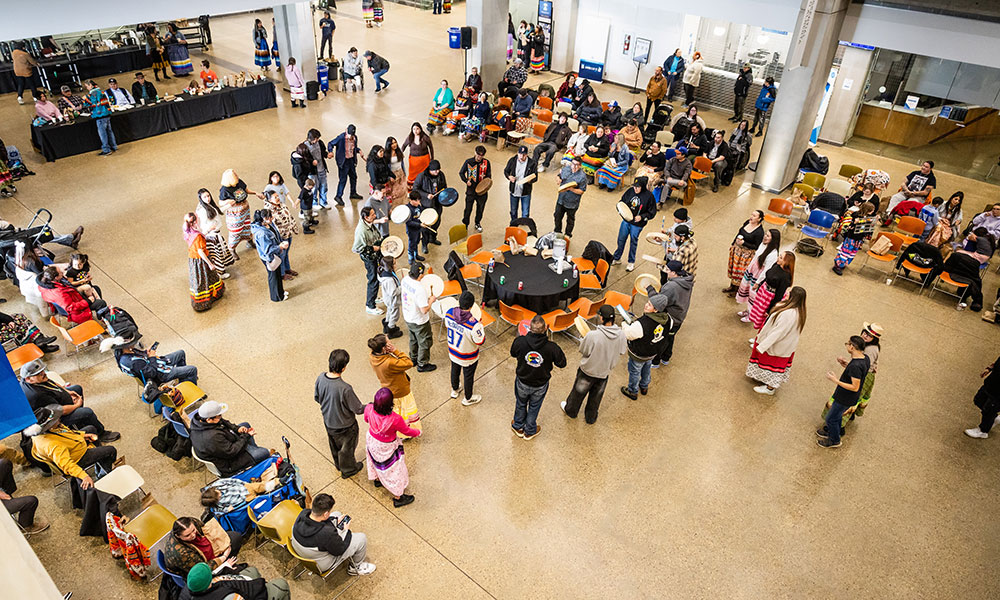 A large group gathers in an open indoor space as people form a round dance circle with drummers at the center.